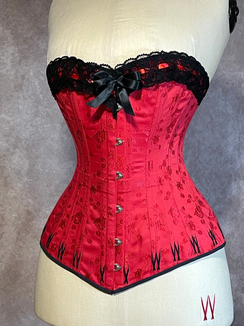 May include: A red corset with black lace trim and a black satin bow. The corset has a floral pattern and is made of a satin-like fabric.