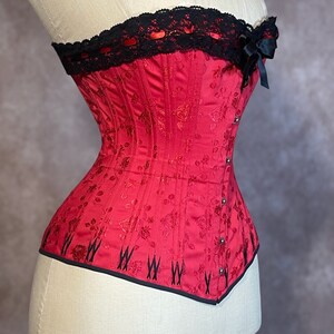 May include: A red corset with black lace trim and a black bow at the top. The corset has a floral pattern and is made of a satin-like fabric.
