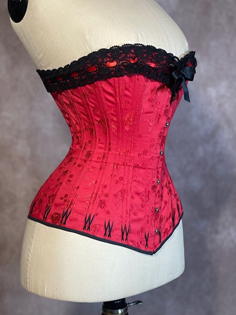 May include: A red corset with black lace trim and a black bow at the top. The corset has a floral pattern and is made of a satin-like fabric.