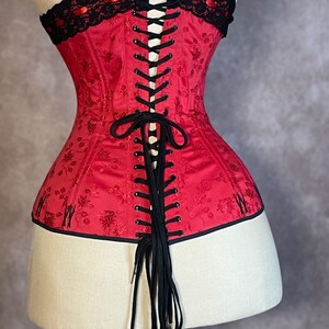 May include: A red corset with black lace trim and black ribbon lacing in the back. The corset has a floral pattern and is made of satin.