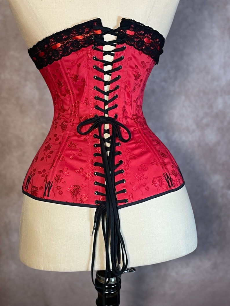 May include: A red corset with black lace trim and black ribbon lacing in the back. The corset has a floral pattern and is made of satin.