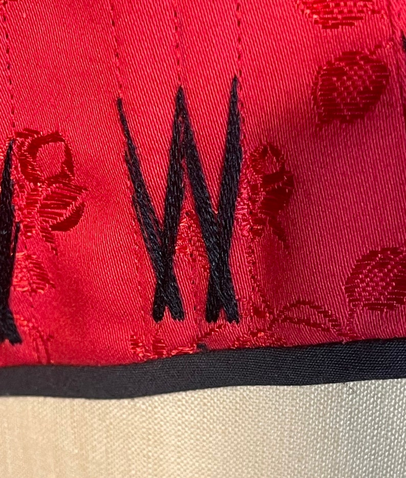 May include: Close-up of a red fabric with a black embroidered "W" and a repeating pattern of red circles. The fabric has a black trim.