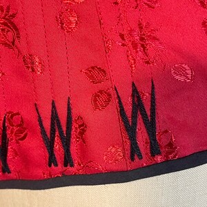May include: Red satin fabric with a floral pattern and black embroidered stitching along the bottom edge.
