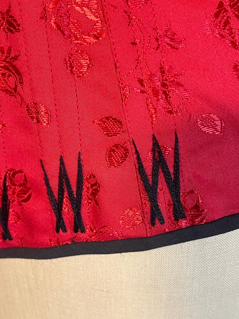 May include: Red satin fabric with a floral pattern and black embroidered stitching along the bottom edge.
