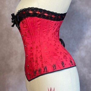 May include: A red corset with black lace trim and a floral pattern. The corset has a boned structure and is designed to cinch the waist.