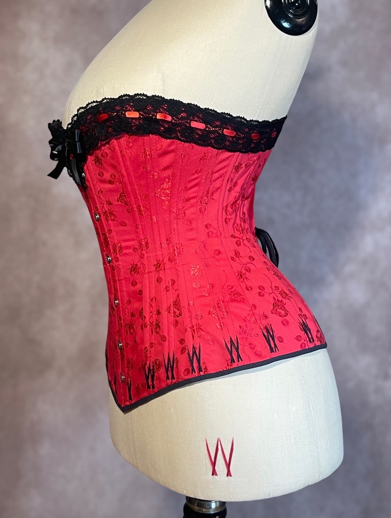 May include: A red corset with black lace trim and a floral pattern. The corset has a boned structure and is designed to cinch the waist.