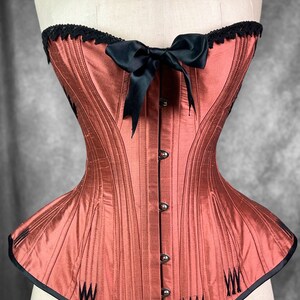 May include: A rust-colored corset with black boning and a black satin bow. The corset has a lace trim at the top and a black trim at the bottom.