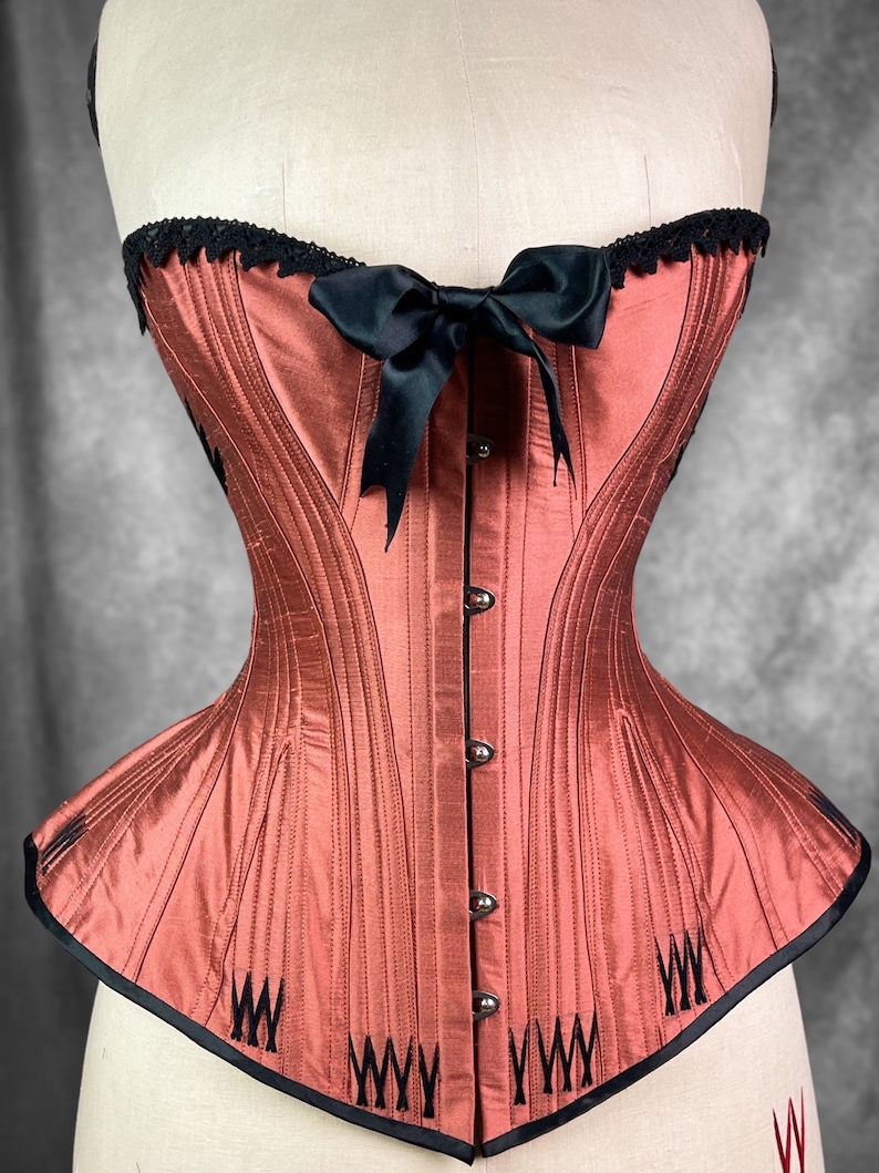 May include: A rust-colored corset with black boning and a black satin bow. The corset has a lace trim at the top and a black trim at the bottom.