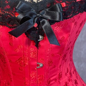 May include: A red corset with a black lace trim and a black satin bow. The corset has a floral pattern and metal clasps.