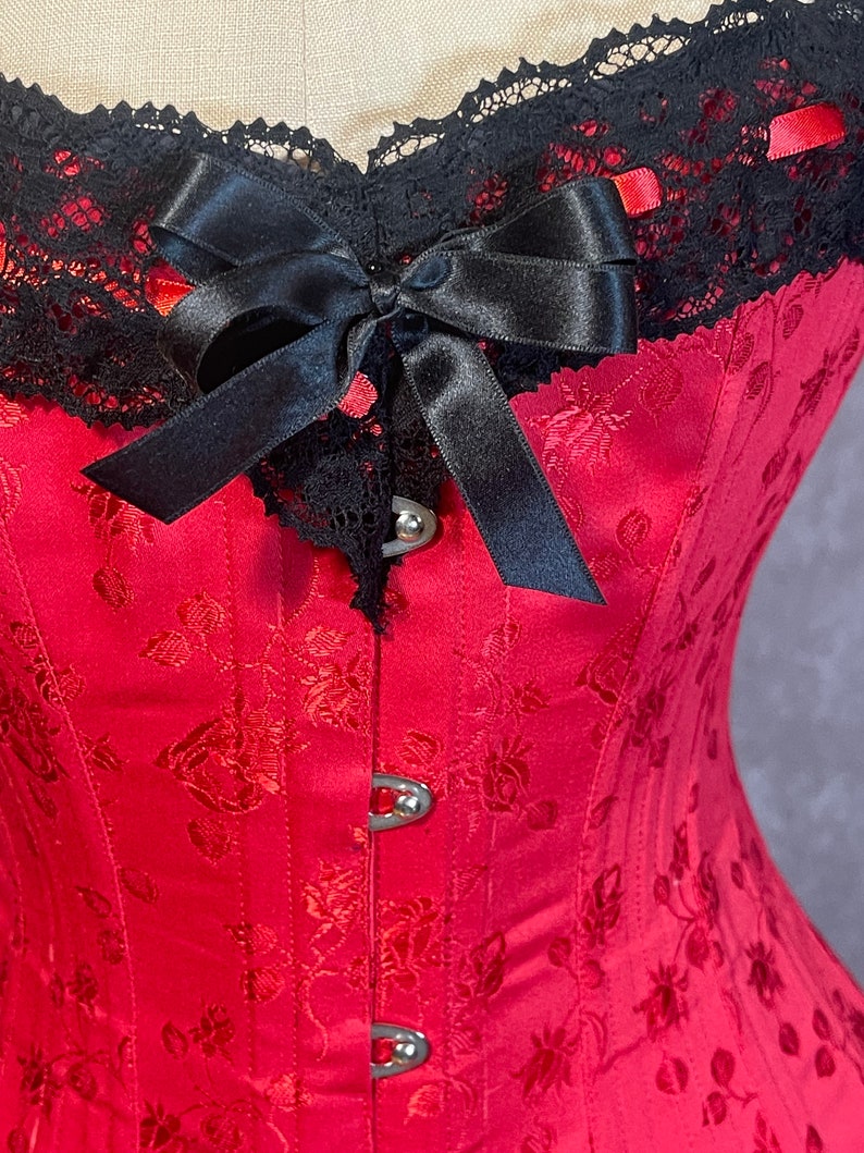 May include: A red corset with a black lace trim and a black satin bow. The corset has a floral pattern and metal clasps.