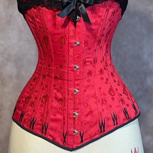 May include: A red corset with a black lace top and a bow. The corset has a floral pattern and is made of satin.