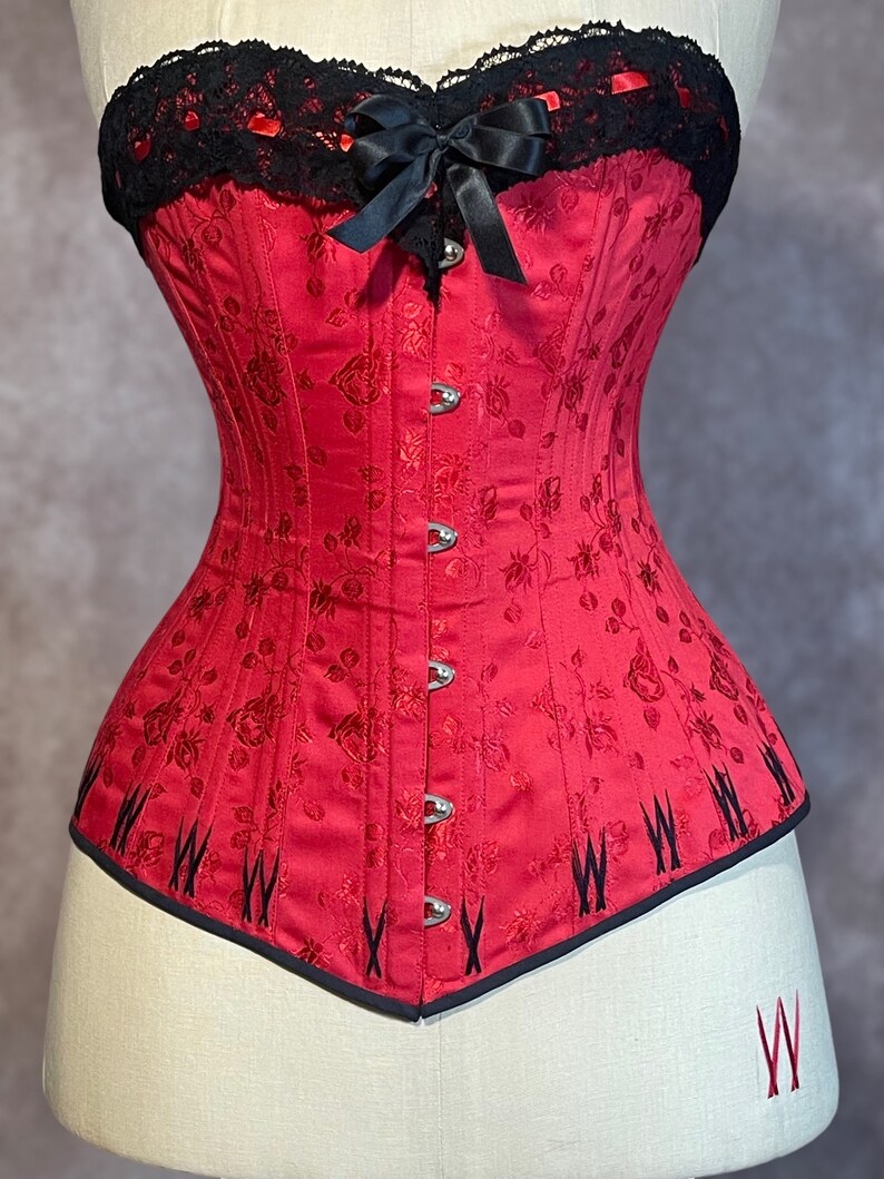 May include: A red corset with a black lace top and a bow. The corset has a floral pattern and is made of satin.