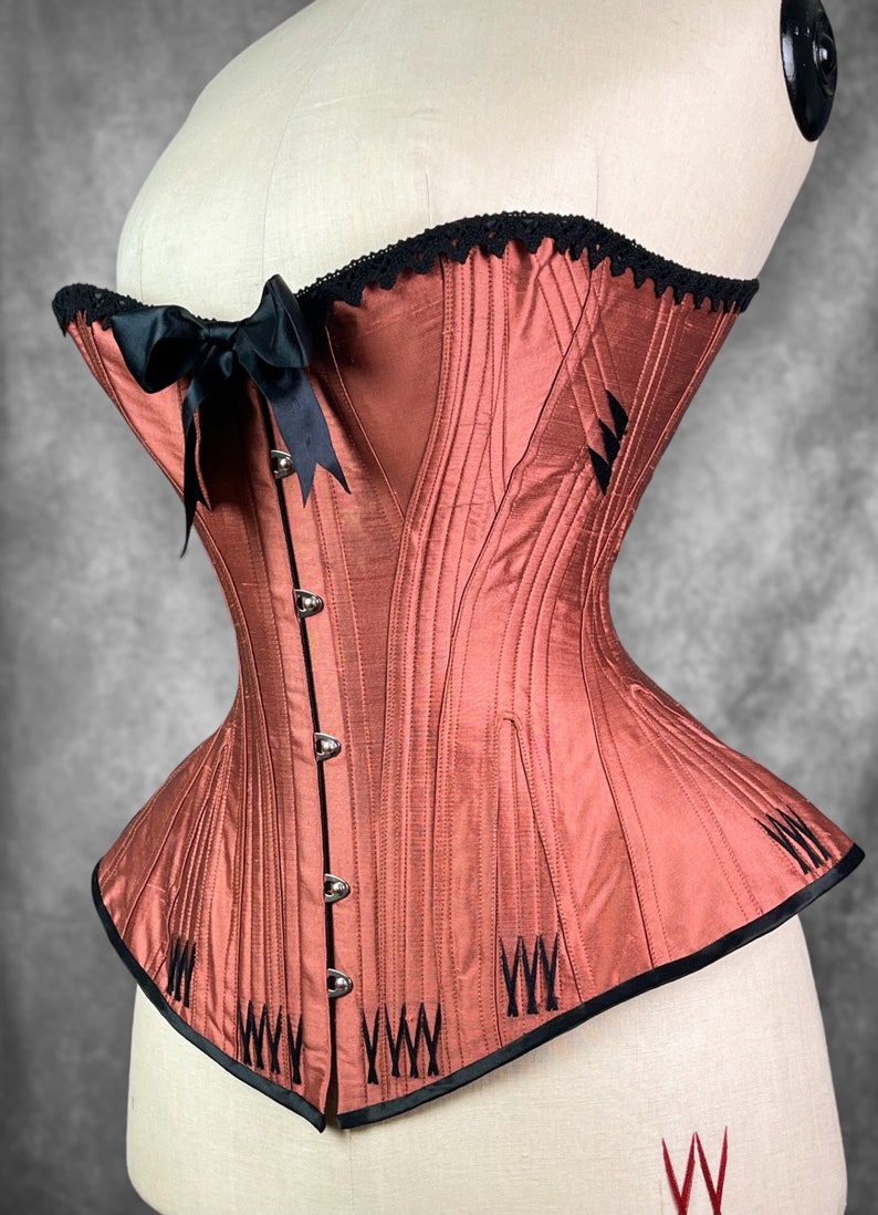 May include: A rust-colored corset with black boning and a black satin ribbon bow. The corset has a lace trim at the top and is made of a smooth, silky fabric.
