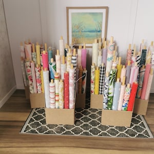 May include: Miniature fabric rolls in diverse patterns and colours, presented in cardboard boxes. The fabrics feature floral, striped, and geometric designs. A framed landscape painting hangs in the background, and a patterned rug is on the floor.