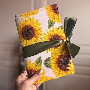 May include: A fabric-covered journal featuring a sunflower pattern with yellow petals and brown centers on a white background. The journal is tied with a green satin ribbon. The edges are finished with a decorative stitch.