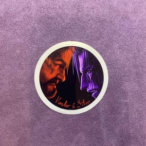 Puede incluir: Una pegatina redonda con un borde blanco y un fondo negro. La pegatina presenta dos caras, una en naranja y rojo y la otra en morado y azul, mirándose. El texto "Vander & Silco" está impreso en blanco en la parte inferior de la pegatina.