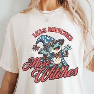 May include: A white t-shirt with a cartoon frog wearing a wizard hat and the text "Less Snitchers More Witches".