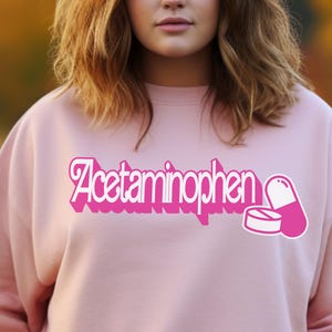 Puede incluir: Una sudadera rosa claro con la palabra "Acetaminophen" en una fuente rosa brillante y estilizada. Un gráfico de pastilla y tableta rosa está a la derecha del texto. La sudadera es de color sólido.