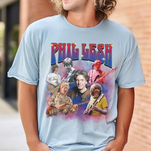 May include: A light blue t-shirt with a vintage-style graphic of Phil Lesh and other musicians. The graphic features the text "Phil Lesh" in a bold, colorful font.