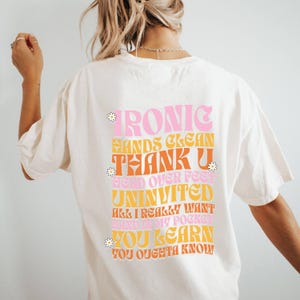 May include: White t-shirt with a retro-style graphic design featuring the text "IRONIC HANDS CLEAN THANK U HEAD OVER FEET UNINVITED ALL I REALLY WANT SAID IN MY POCKET YOU LEARN YOU OUGHTA KNOW" in pink, orange, and yellow.