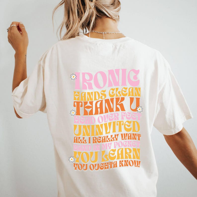 May include: White t-shirt with a retro-style graphic design featuring the text "IRONIC HANDS CLEAN THANK U HEAD OVER FEET UNINVITED ALL I REALLY WANT SAID IN MY POCKET YOU LEARN YOU OUGHTA KNOW" in pink, orange, and yellow.