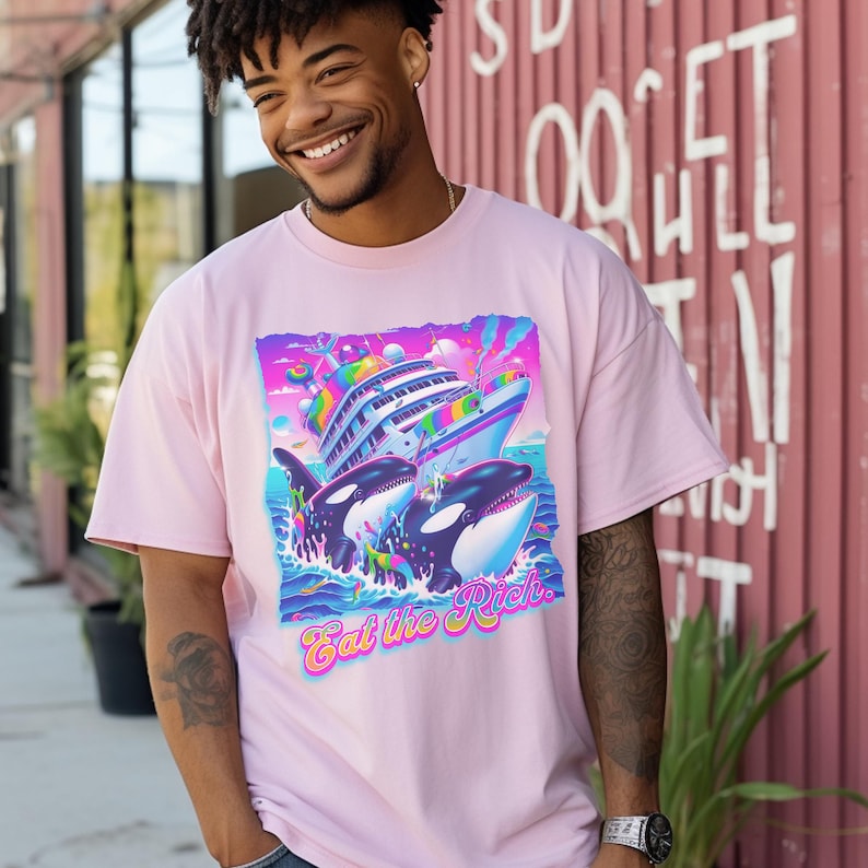 May include: A light pink t-shirt featuring a vibrant graphic of a cruise ship, two orcas, and ocean waves. The text "Eat the Rich" is displayed in a colorful, stylized font. The shirt is made of a soft material.