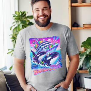 May include: A heather grey t-shirt with a graphic of two orcas jumping out of the water and attacking a large yacht. The graphic is in a bright, colorful style with rainbow accents. The text "Eat the Rich" is printed below the graphic.