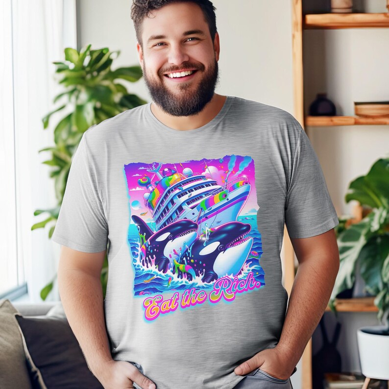 May include: A heather grey t-shirt with a graphic of two orcas jumping out of the water and attacking a large yacht. The graphic is in a bright, colorful style with rainbow accents. The text "Eat the Rich" is printed below the graphic.