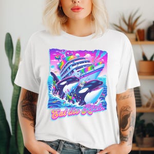May include: A white t-shirt with a graphic of two orcas swimming towards a large, colorful cruise ship. The graphic is surrounded by a rainbow of colors and the text "Eat the Rich." is printed below the graphic.