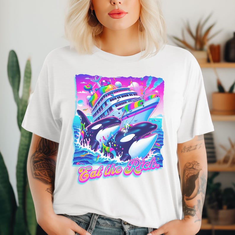 May include: A white t-shirt with a graphic of two orcas swimming towards a large, colorful cruise ship. The graphic is surrounded by a rainbow of colors and the text "Eat the Rich." is printed below the graphic.