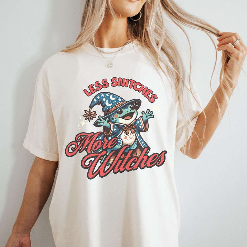 May include: A white t-shirt with a cartoon frog wearing a wizard hat and the text "Less Snitchers More Witches".