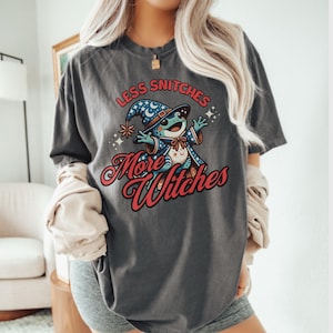May include: A dark gray t-shirt with a graphic of a frog wearing a wizard hat and robe. The text "Less Snitchers More Witches" is printed in red on the shirt.