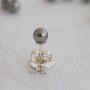 May include: A silver earring with a small, round, dark grey bead. The earring is on a white surface with other dark grey beads scattered around it.