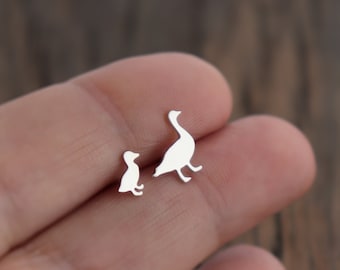 Op de afbeelding: Een paar zilverkleurige oorknopjes met de silhouetten van een klein eendenkuiken en een volwassen gans. De oorbellen hebben een gladde, metalen afwerking en zijn ontworpen als individuele vogelvormen, elk voorzien van een stekertje.