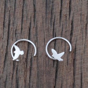 May include: Two silver earrings, each with a bird silhouette inside a curved shape. The earrings are set against a dark brown, textured wooden background. The bird design gives the impression of flight.