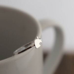 May include: A silver ring with a leaf-shaped charm. The ring is thin and delicate, with the leaf charm positioned on the top. The ring is shown against a white background, with a white mug in the background.