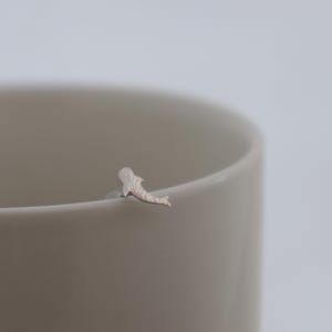 May include: A small, silver-toned stud earring shaped like a whale shark, featuring a textured surface and a clear plastic backing, resting on the rim of a smooth, light-colored ceramic mug. The earring is metallic with a matte finish.