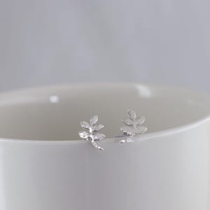 May include: A pair of silver leaf stud earrings. The earrings are small and delicate, with a textured surface. The leaves have a detailed vein pattern. The earrings are displayed on a white surface.
