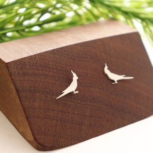 May include: A pair of silver-toned stud earrings shaped like cockatiel birds. Each earring features a detailed silhouette of a bird with a prominent crest, a pointed beak, and a long tail, rendered in a smooth, metallic finish with a polished surface.
