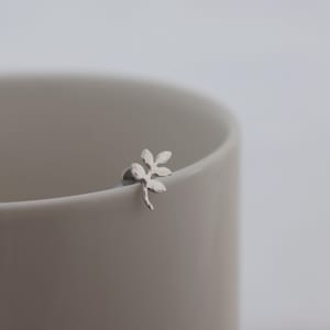 May include: A small silver-toned stud earring shaped like a delicate sprig with three distinct leaves. The earring features a textured metallic finish and is positioned on the curved rim of a smooth, light-coloured ceramic cup.