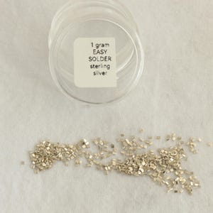 Può includere: Un contenitore di plastica trasparente con un'etichetta che recita "1 gram EASY SOLDER sterling silver" è posto su una superficie bianca. Sotto, piccoli pezzi di saldatura argentata sono sparsi, per gioielli e artigianato.