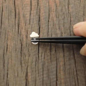 May include: A pair of black tweezers holding a small silver charm in the shape of a cloud with a raindrop inside. The charm is on a wooden surface.