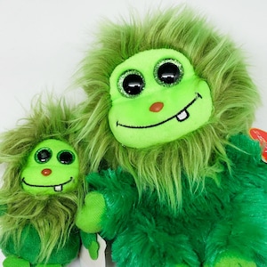 May include: Two green plush toys with googly eyes and toothy grins. The larger toy is sitting with its legs crossed, while the smaller toy is standing. Both toys have a fuzzy texture.