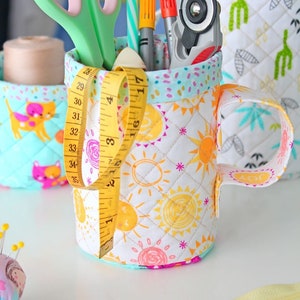 May include: A turquoise fabric mug with a yellow measuring tape, scissors, markers, and a straw. The mug has a white background with a pattern of suns and stars. The mug is sitting on a white surface.