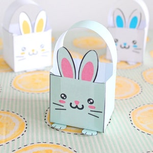May include: Three paper bunny gift bags with handles. The bags are white, light blue, and light green. The bunny faces are drawn on the front of the bags. The bags are sitting on a green and white striped surface with yellow lemon slices.