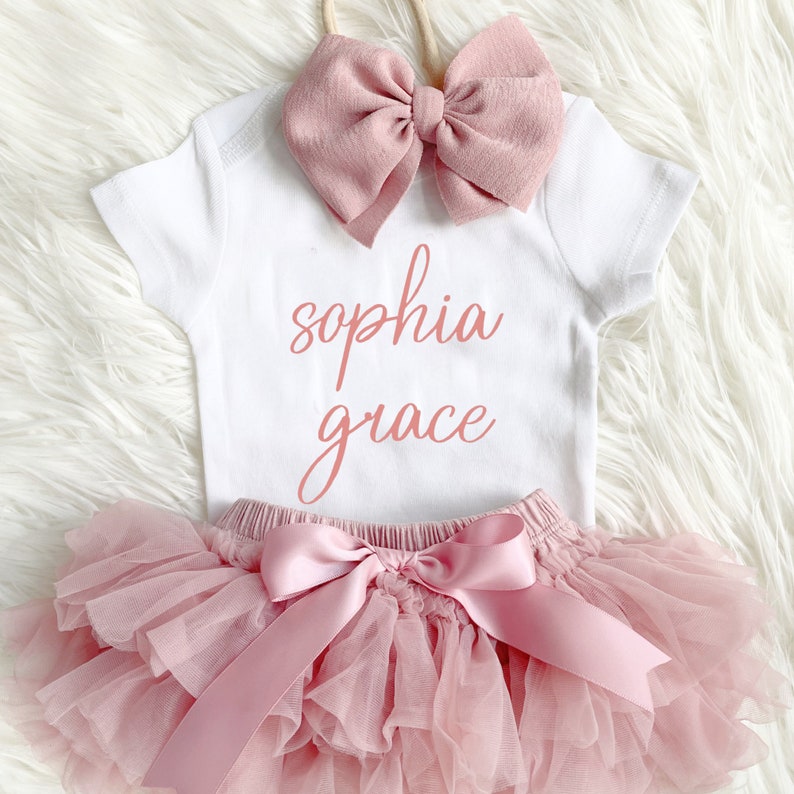 Puede incluir: Un body blanco para beb&eacute; con el texto "sophia grace" en escritura rosa. El body se combina con una falda de tul rosa y una diadema con lazo rosa.