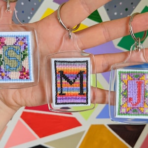 Può includere: Tre portachiavi in acrilico trasparente con iniziali ricamate a punto croce. Le iniziali sono "S", "M" e "J", ciascuna con una diversa combinazione di colori. La "S" è circondata da fiori, la "M" ha un gradiente arcobaleno e la "J" ha uno sfondo viola.