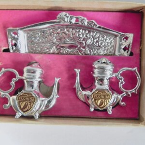 May include: A vintage souvenir set featuring a silver-toned metal napkin holder and two teapot-shaped salt and pepper shakers. The shakers have a gold-toned emblem that reads "FLORIDA". The set is displayed on a pink background inside a cardboard box.