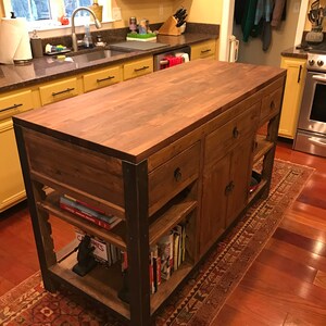 Pu&ograve; includere: Un'isola da cucina in legno con un piano di lavoro in legno di macellaio, mensole aperte e cassetti. L'isola ha una struttura in metallo nero e si trova su un tappeto rosso e marrone con motivo.
