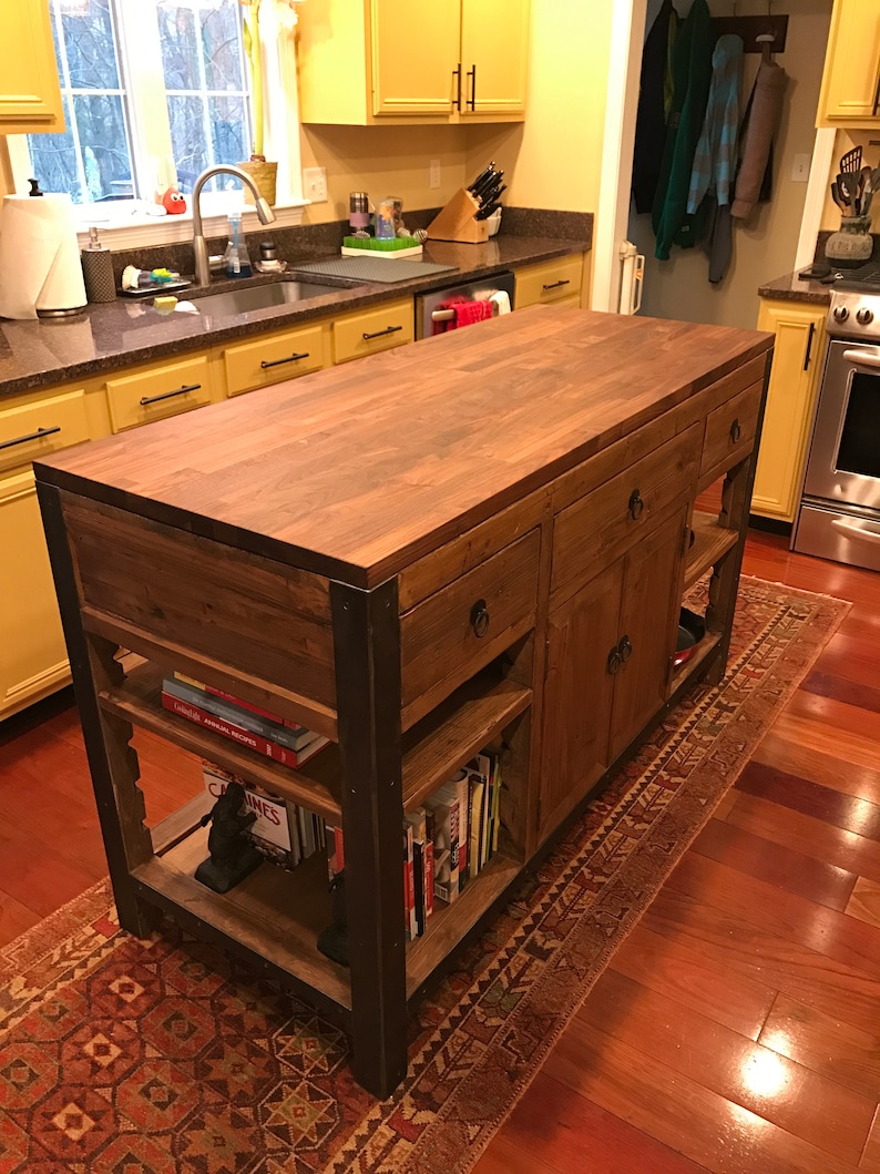 Pu&ograve; includere: Un'isola da cucina in legno con un piano di lavoro in legno di macellaio, mensole aperte e cassetti. L'isola ha una struttura in metallo nero e si trova su un tappeto rosso e marrone con motivo.
